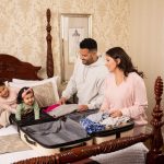 a man and woman standing next to a hotel bed with an open suitcase full of clothing