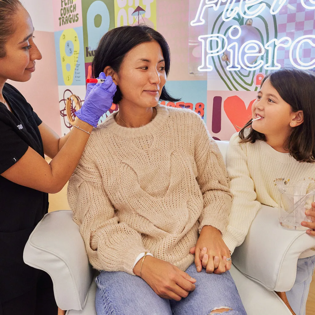 mother and daughter holding hands while mother gets ears pierced by nurse